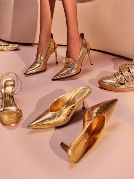 Imagem mostra alguns calçados dourados pelo chão, são peças da coleção de dia das mães da Santa Lolla. Scarpin mule, sandália de salto fino e tênis.